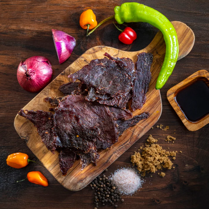 A wooden cutting board displaying pieces of jerky, surrounded by colorful vegetables and seasonings, including peppers, onions, salt, sugar, and a small bowl of dark sauce.